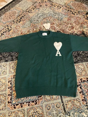 Grön  tröja från Ami Paris - Mörkgrön stickad tröja från Ami Paris med rund halsringning och ribbade muddar. På bröstet finns ett vitt hjärta och bokstaven 'A' som är typiskt för märket. Perfekt för dig som gillar stilrena detaljer och vill ha något unikt i garderoben.