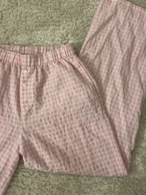Rosa rutiga pyjamasbyxor - Säljer ett par mysiga pyjamasbyxor i ljusrosa och vitt rutigt mönster. Byxorna har resår i midjan och tre vita knappar framtill. Materialet är mjukt och luftigt💕