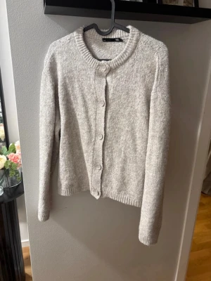 Ljusgrå/ beige kofta med knappar - Mysig ljusgrå kofta med rund halsringning och knäppning framtill. Koftan har långa ärmar och ribbade muddar vid ärmslut och nederkant. Perfekt för lager på lager och enkel att matcha med olika outfits.