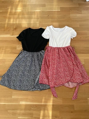 Två blommiga kortklänningar - Säljer två söta kortklänningar med blommigt mönster. Ena har svart överdel och blåvit kjol, den andra har vit överdel och rödvit kjol med knytband i midjan. Båda har rund halsringning och korta ärmar. Perfekta för sommarens alla tillfällen!