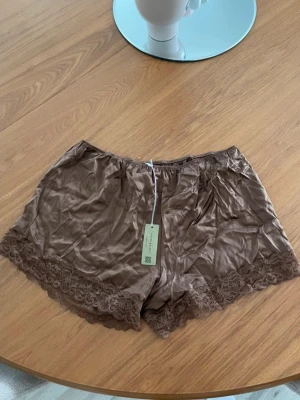 Bruna pyjamasshorts med spetsdetaljer - Snygga bruna pyjamasshorts i satin med fina spetsdetaljer längs bensluten. Shortsen har resår i midjan för extra komfort och en mjuk, glansig finish. Finns på fler sidor