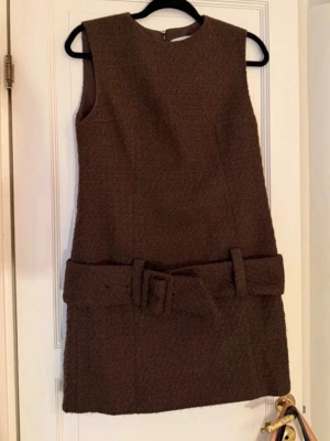 Brun ärmlös klänning med bälte - Snygg brun kortklänning i grovt tyg med bredt matchande bälte i midjan. Klänningen är ärmlös och har en enkel, rak passform som ger en stilren look. Perfekt för dig som gillar retrovibbar och vill ha något unikt i garderoben. Aldrig använd!