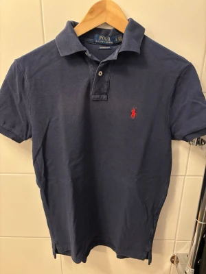 Ralph Lauren Pike - Klassisk marinblå pikétröja från Ralph Lauren med röd broderad logga på bröstet. Modellen är Custom Slim Fit med korta ärmar, krage och knappar framtill. Perfekt för en stilren och sportig look. Skick 8/10✅