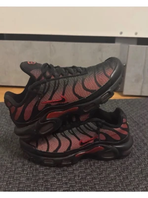 Nike Air Max Plus TN Svart/Röd - Nike Air Max Plus TN sneakers i svart och rött med ikoniska vågmönster på ovandelen. Skorna har en mesh-överdel, svarta och röda detaljer samt synlig Air Max-dämpning i sulan. Röda Swoosh-loggor på sidorna och TN-logga på hälen. Perfekta för dig som gillar streetwear.