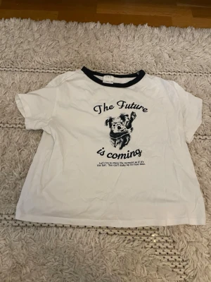 Vit t-shirt med tryck från Zara - Vit t-shirt från Zara med svart text 'The Future is coming' och en koala på bröstet. Svart ribbad kant runt halsen och korta ärmar. Perfekt för en avslappnad och cool stil.