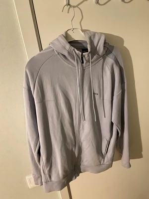 Ljusblå hoodie från BOSS - Säljer en stilren ljusblå hoodie från BOSS med dragkedja framtill och justerbar huva med snören. Diskret BOSS-logga på bröstet och två fickor på sidorna. Perfekt för en avslappnad och trendig look.
