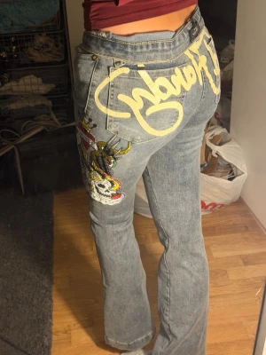 Blå Ed Hardy jeans med tryck - Unika blå jeans från Ed Hardy med stort gult graffiti-tryck på bakfickan och färgglad tatuering-inspirerad design på benet. Jeansen har normal passform och är tillverkade i klassiskt denim-material. Perfekta för dig som vill sticka ut med coola detaljer. Dem är mer åt de mid rise hållet men kan enkelt vikas ner, jätte jätte stretchiga men fortfarande jeans material så jätte sköna på❤️har aldrig använt dem men drog tyvärr bort lappen endå💔 köpte på ed Hardy butiken i usa!! Pris kan diskuteras❤️