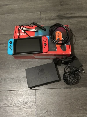 Nintendo Switch med andra tillbehör  - Nintendo Switch med blå och röd Joy-Con, originalförpackning, laddare, dockningsstation. Konsolen och tillbehören ser ut att vara i gott skick utan synligt slitage.