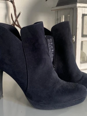 Tamaris Svarta mockastövletter med klack - Snygga svarta stövletter i mocka med hög klack och dragkedja på sidan. Skorna har en stilren design med rundad tå och diskret plattform. Perfekta för dig som vill ha en trendig och elegant look.