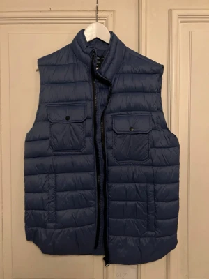 Mörkblå dunväst med fickor - Snygg mörkblå dunväst med hög krage och två stora bröstfickor med lock och knapp. Västen har quiltad design och dragkedja framtill. Perfekt för lager-på-lager och ger en clean look till din outfit.