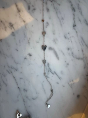 Silverfärgat armband med hjärtan - Snyggt armband i silverfärgad metall med fem små hjärtan längs kedjan. Enkel och stilren design som passar till många olika looks. Justerbar längd med liten bricka vid låset. Perfekt accessoar för dig som gillar subtila detaljer.