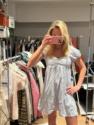 Blåvit randig babydollklänning - Söt babydollklänning med blå och vita ränder. Klänningen har korta puffärmar, markerad underbyst och en luftig, A-linjeformad passform. Perfekt för en avslappnad och trendig look. Materialet ser ut att vara bomull eller bomullsblandning.