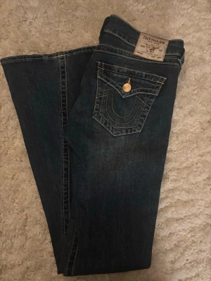 True Religion Joey Low Rise Flare Jeans - Mörkblå jeans från True Religion, modell Joey Low Rise Flare. Klassisk femficksdesign med dekorativa sömmar och guldfärgad knapp. Snyggt broderad bakficka och låg midja med utsvängda ben. Perfekta för en trendig och avslappnad stil.