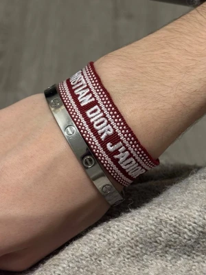 Christian Dior J'Adior armband vinröd - Snyggt vävt armband från Christian Dior i vinrött och vitt med texten 'J'ADIOR'. Armbandet har justerbar knytning med tofsar i ändarna. Perfekt accessoar för att lyfta din stil med en lyxig touch.