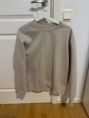 Beige basic sweatshirt unisex - En stilren beige sweatshirt med rund hals och ribbade muddar vid ärmslut och nederkant. Tröjan har en avslappnad passform och är tillverkad i mjukt material, perfekt för en casual look.