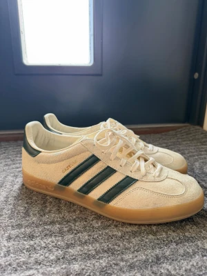 Adidas Gazelle beige/gröna sneakers - Snygga Adidas Gazelle sneakers i beige mocka med mörkgröna läderdetaljer och klassiska tre ränder på sidan. Skorna har gummisula i brunt och vita skosnören. Perfekt för dig som gillar retrostil och vill ha något stilrent till vardagslooken. Knappt använda 1-2 ggr. Nypris 3,6k pga de sällsynt färg att hitta