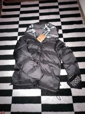 Svart pufferjacka från Burberry - Snygg svart pufferjacka från Burberry med klassiskt rutigt foder i huvan. Jackan har dragkedja och knappar framtill, två sidofickor och justerbar nederkant. Perfekt för kalla vinterdagar och riktigt trendig look.