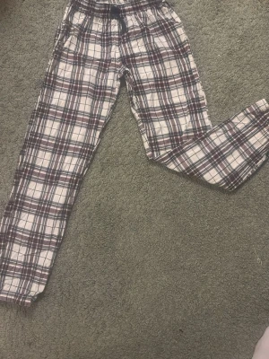 Rutiga pyjamasbyxor i bomull - Mysiga pyjamasbyxor med rutigt mönster i vitt, vinrött och mörkblått. Tillverkade i mjuk bomull med avslappnad passform och resår i midjan. Perfekta för chillkvällar hemma.