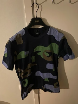 Camouflage t-shirt från BOSS - Säljer en snygg camouflage t-shirt från BOSS med korta ärmar. T-shirten har ett klassiskt camomönster i grönt, blått, svart och brunt. Perfekt för dig som gillar streetwear och vill sticka ut lite extra.