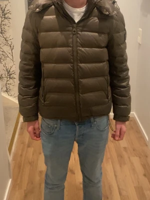 Mörkgrön dunjacka med huva - Säljer en mörkgrön dunjacka med huva och dragkedja framtill. Jackan har en puffig design med horisontella sömmar och två sidofickor. Perfekt för kalla vinterdagar och riktigt skön att bära. Passar dig som vill ha en stilren och varm jacka. Pris kan diskuteras!