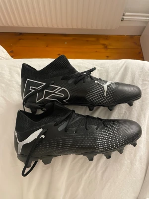 Svarta Puma Future fotbollsskor - Säljer ett par svarta Puma Future fotbollsskor med vita detaljer och logga. Skorna har en strumpliknande krage för extra stöd runt ankeln och snörning för bra passform. Ovandelen är i syntetmaterial med textur för bättre bollkontroll. Perfekta för spel på gräsplan.