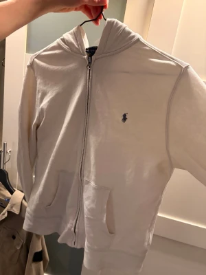 Vit hoodie från Polo Ralph Lauren - Snygg vit hoodie med dragkedja från Polo Ralph Lauren. Klassisk design med två fickor fram, huva och den ikoniska blå loggan broderad på bröstet. Tillverkad i mjukt bomullsmaterial som är skönt att ha på sig. Perfekt för en avslappnad och stilren look. (14-16 år)
