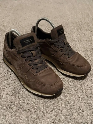 Bruna sneakers från TOD'S i mocka - Snygga bruna sneakers från TOD'S i mjuk mocka med mörkbruna skosnören och svart logotyp på plösen. Skorna har en klassisk låg modell och en vit sula med svarta detaljer. Perfekta för en stilren och avslappnad look.