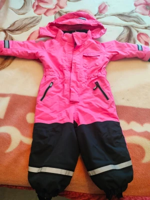 Rosa och svarta täckbyxor med huva - Täckbyxor i overallmodell med starkt rosa överdel och svarta ben, reflexdetaljer vid benslut och dragkedjeförsedda fickor. Overallen har huva och resår i midjan för skön passform. Materialet är slitstarkt och vattenavvisande, perfekt för kalla dagar.