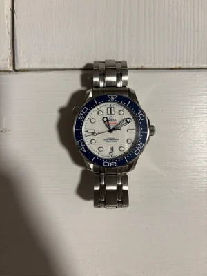 Omega Seamaster klocka blå/vit stål - Säljer en Omega Seamaster klocka med blå vridring och vit urtavla. Klockan har datumvisning, självlysande index och visare samt robust länkarmband i rostfritt stål. Baksidan har genomskinligt fönster som visar urverket. Perfekt för dig som gillar klassisk och sportig stil.