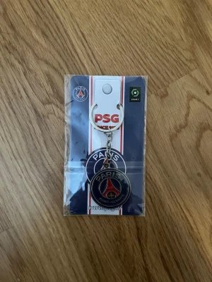 PSG nyckelring Paris Saint-Germain - Snygg PSG nyckelring i metall med klubbens logga i blått, rött och vitt. Perfekt accessoar för dig som älskar fotboll och vill visa ditt stöd för Paris Saint-Germain. Rund form och robust kedja.