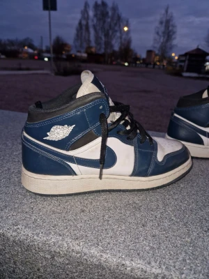 Nike Air Jordan 1 Mid blå/vit - Snygga Nike Air Jordan 1 Mid sneakers i blått och vitt läder med svarta detaljer och klassisk Jordan-logga på sidan. Skorna har svart snörning och vit sula. Perfekta för dig som gillar streetwear och vill sticka ut med en ikonisk modell.