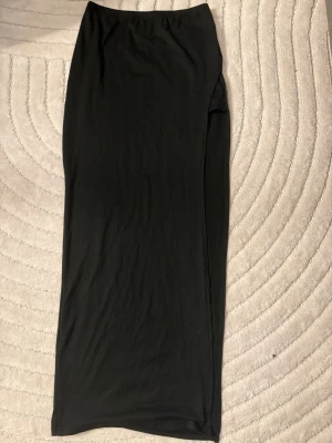 Svart långkjol med slits - En svart långkjol i mjukt material med en slits på sidan. Kjolen har en enkel och stilren design som passar till många olika looks. Perfekt för dig som gillar minimalistisk stil.