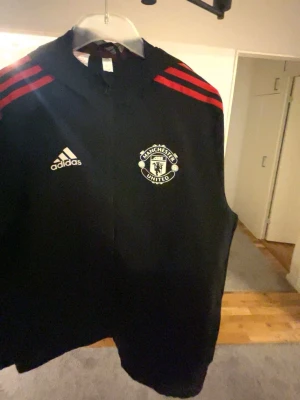 Manchester United sportkofta från Adidas - Snygg svart hoodie från Adidas med Manchester Uniteds klubbmärke på bröstet. Tröjan har röda ränder på axlarna och Adidas-logga i vitt. Perfekt för dig som älskar fotboll och vill representera ditt lag med stil.