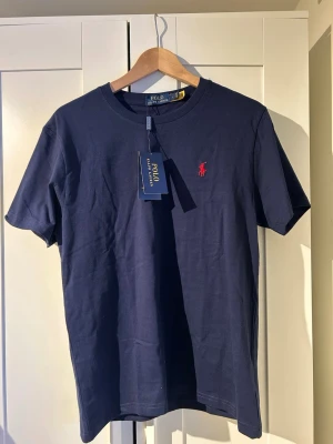 Marinblå t-shirt från Polo Ralph Lauren - Klassisk marinblå t-shirt från Polo Ralph Lauren med röd broderad logga på bröstet. Tillverkad i mjuk bomull och har en normal passform med korta ärmar och rund hals. Perfekt basplagg för en clean och stilren look. Helt ny priset kan diskuteras 