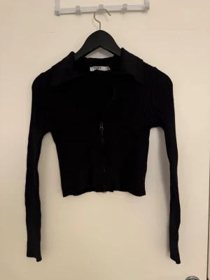 Svart ribbad ziptröja från Nakd - Snygg svart ribbad ziptröja med krage och långa ärmar. Tröjan är croppad och har dragkedja framtill, vilket ger en trendig och stilren look. Perfekt att matcha med höga jeans eller kjol.