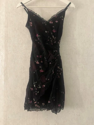 Svart blommig klänning - Säljer en svart kortklänning med tunna axelband.  Klänningen har ett blommigt mönster i rosa och grönt samt volangkant nedtill. Perfekt för dig som gillar romantiska detaljer och en feminin look.