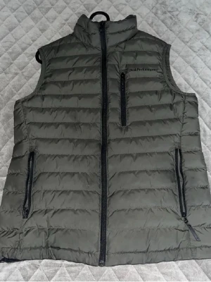 Olivgrön dunväst Peak Performance - Snygg olivgrön dunväst från Peak Performance med hög krage och dragkedja framtill. Västen har två sidofickor med dragkedja och en bröstficka. Quiltad design och lätt vadderad för extra värme. Perfekt lager på lager-plagg för kyliga dagar.