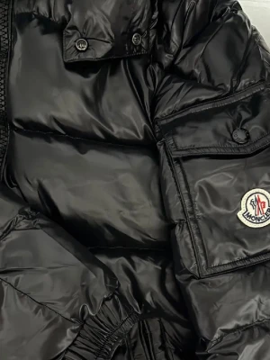  Moncler Maya  - Snygg svart dunjacka från Moncler med glansig finish och klassisk logotyp på ärmen. Jackan har dragkedja framtill, fickor med lock och knapp samt resår i midjan för en schysst passform. Perfekt för kalla vinterdagar.