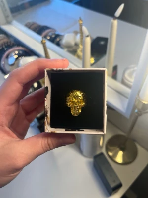 Guldfärgad ring med dödskalle - Cool ring i guldfärgad metall med en detaljerad dödskalle på framsidan. Ringen har en chunky design och glänsande finish som verkligen sticker ut. Perfekt för dig som gillar edgy accessoarer och vill göra ett statement.