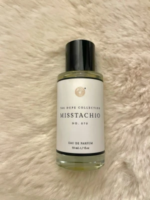 Curiosa Misstachio - Säljer en stilren parfym i genomskinlig glasflaska med svart kork. Parfymen har en ljusgul vätska och en enkel, modern design med vit etikett. Perfekt för dig som gillar minimalistiska doftflaskor.