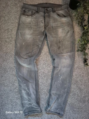 Dsquared2, Grå slim fit jeans med slitningar - Säljer ett par grå slim jeans från dsquared2 med coola slitningar och vita färgstänk för en trendig, avslappnad look. Jeansen har klassisk femficksmodell och knappgylf. Perfekta för dig som gillar streetstyle och vill sticka ut lite extra.