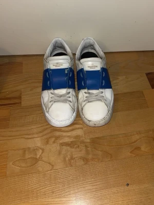 Valentino vita sneakers med blå detalj - Snygga vita sneakers från Valentino med bred blå läderdetalj över snörningen. Klassisk låg modell med rund tå och vita skosnören. Tillverkade i läder för en exklusiv känsla och stilren look.