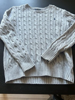 Ljusgrå kabelstickad tröja Polo Ralph Lauren - Snygg ljusgrå kabelstickad tröja från Polo Ralph Lauren. Tröjan har rund halsringning, långa ärmar och ribbade muddar. Klassisk liten broderad logga på bröstet.