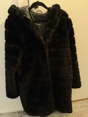 Svart fuskpälsjacka från Amisu - Säljer en svart fuskpälsjacka från Amisu med huva och mjukt foder. Jackan har en oversized passform och breda, horisontella paneler som ger en lyxig look. Perfekt för kalla dagar när du vill vara både varm och snygg.