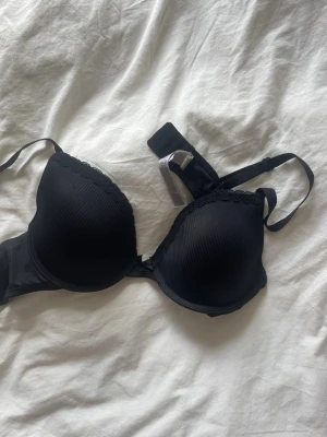 Svart push-up bh med spetskant - Snygg svart push-up bh med lätt vadderade kupor och dekorativ spetskant upptill. Justerbara axelband och hakknäppning bak. Materialet är slätt och stretchigt för skön passform. Perfekt för dig som vill ha lite extra lyft och en klassisk look.