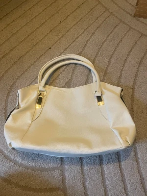 Vit handväska med gulddetaljer - Snygg vit handväska i konstläder med guldfärgade metalldetaljer och dubbla handtag. Väskan har en avtagbar axelrem som gör den flexibel att bära. Stilren och rymlig modell som passar till många olika outfits. Se 6e bilden fiins en liten skada som syns knappt