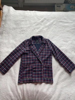 Rutig kavaj från Monki - Snygg rutig kavaj från Monki i blått, rött och vitt. Dubbelknäppt modell med två stora fickor framtill och klassisk krage. Kavajen har helfodrad insida i mörkblått tyg. Storlek XS.