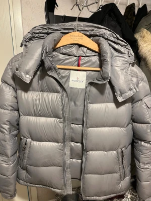 Grå Moncler maya   - Snygg grå dunjacka från Moncler Maya kollektionen med klassisk pufferdesign och avtagbar huva. Jackan har dragkedja framtill, två fickor med dragkedja och Moncler-logga på ärmen. Perfekt för kalla dagar och riktigt trendig look.