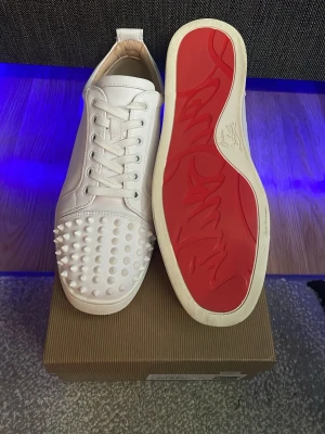 Vita Louboutin sneakers med nitar - Säljer ett par vita sneakers från Christian Louboutin med ikoniska röda sulor och coola nitar på tån. Skorna är i läder och har vita skosnören. Perfekta för dig som vill sticka ut med en edgy och lyxig stil.