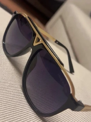 Lv solglasögon  - Snygga solglasögon från Louis Vuitton med svart båge och guldiga detaljer. Glasen är mörka och har en klassisk aviatorform. Perfekt accessoar för att lyfta din stil med en lyxig touch.